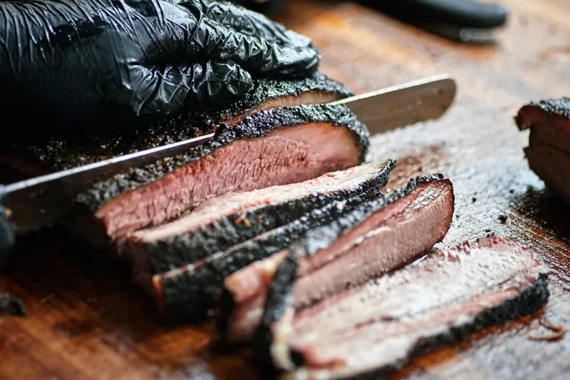 Gloved pitmaster slicing a peppery, bark-crusted smoked brisket on a wooden board at ZZQ Texas Craft Barbeque, Richmond