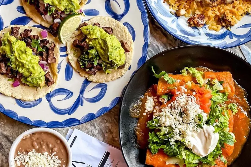A spread at La Santa Modern Mexican in Raleigh: carne asada street tacos with guacamole on a blue Talavera plate, a chicken-and-rice plate, enchiladas, and refried beans beside a La Santa menu card