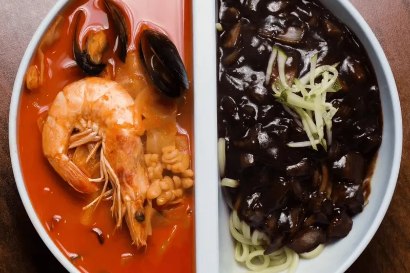 Split bowl of jjamppong spicy seafood noodle soup with shrimp and mussels beside black-bean jjajangmyeon noodles at Man Chun Hong