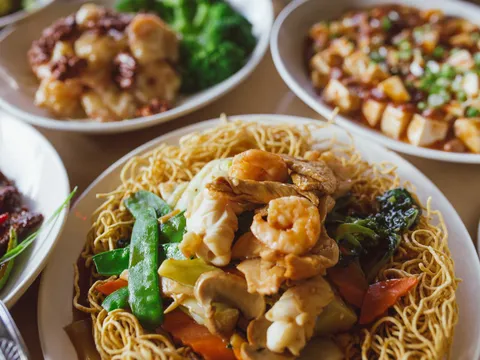 A spread of Dumpling Inn dishes on a wood table — a crispy pan-fried noodle plate topped with shrimp, chicken and vegetables in the foreground, with walnut shrimp and mapo tofu behind
