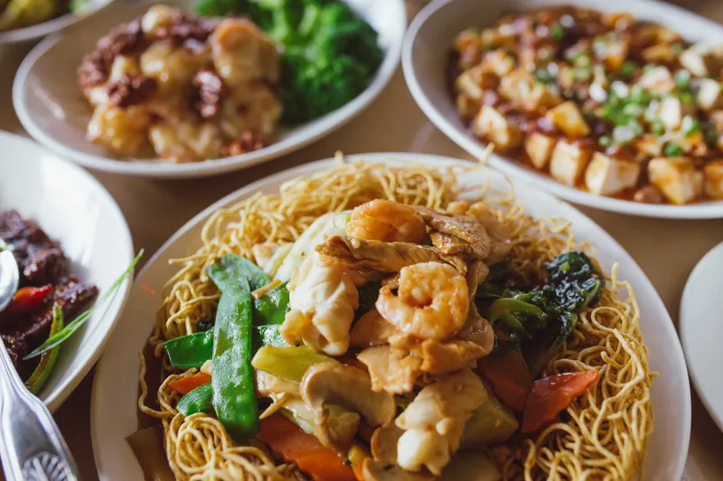 A spread of Dumpling Inn dishes on a wood table — a crispy pan-fried noodle plate topped with shrimp, chicken and vegetables in the foreground, with walnut shrimp and mapo tofu behind