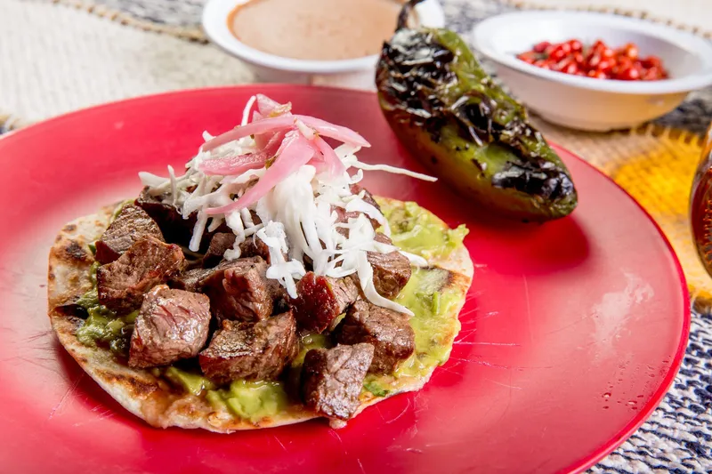 Mesquite-grilled carne asada taco on a red plate with guacamole, pickled onion and cabbage, a charred poblano and refried beans, at Casa Asada Taqueria y Cerveceria in Tucson