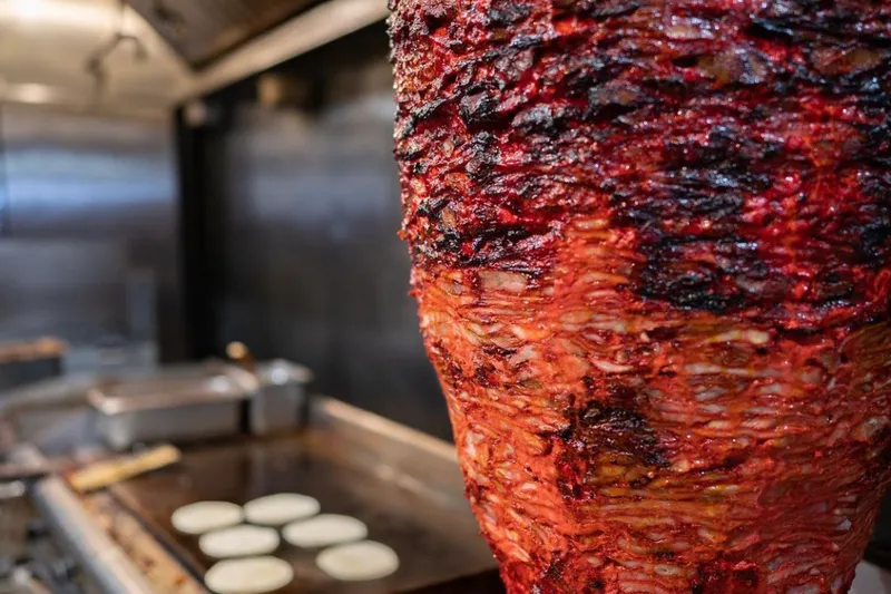 Marinated al pastor pork (trompo) cooking on a vertical spit beside a griddle of tortillas at Trompo Taqueria & Burgers