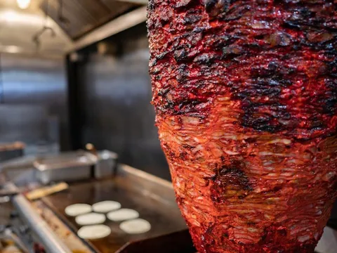 Marinated al pastor pork (trompo) cooking on a vertical spit beside a griddle of tortillas at Trompo Taqueria & Burgers