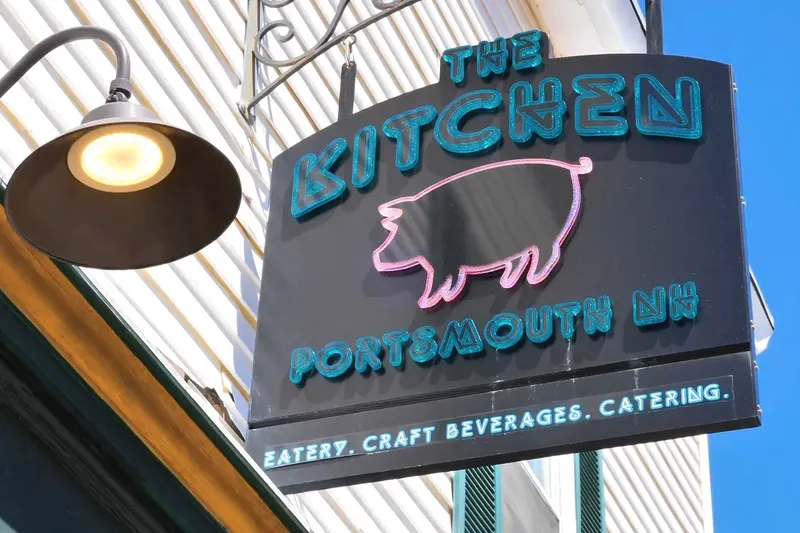 Painted sign reading The Kitchen, Portsmouth NH, with a pig motif, lit by a gooseneck lamp