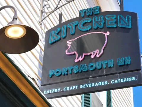 Painted sign reading The Kitchen, Portsmouth NH, with a pig motif, lit by a gooseneck lamp