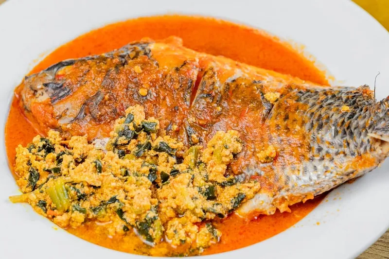 Whole fish simmered in red pepper sauce with egusi (ground melon seed and greens) on a white plate at Buka, Brooklyn