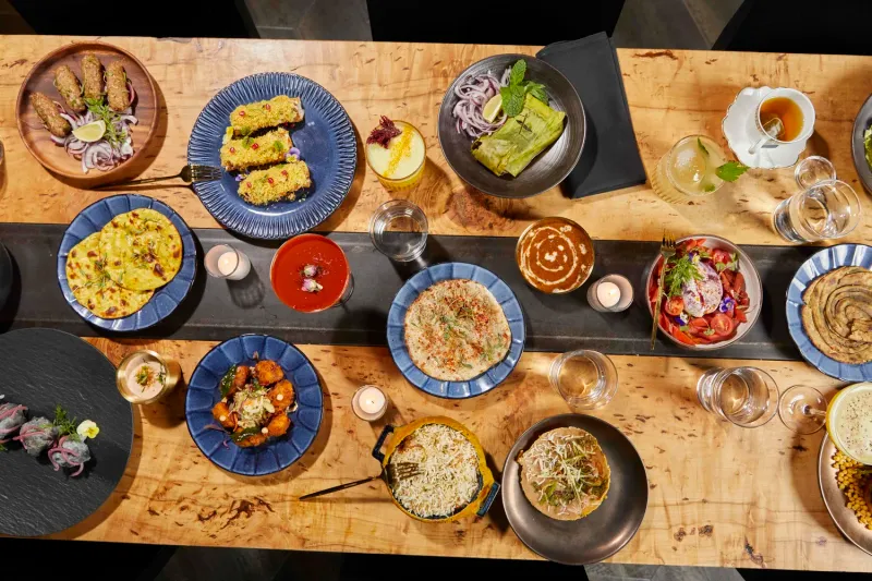 An overhead spread of Kricket Club dishes — seekh kebabs, breads, curries, biryani, salad and cocktails — arranged on a wood table