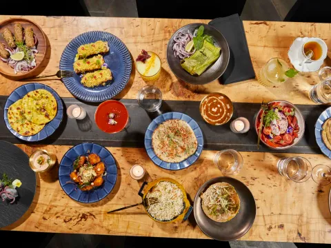 An overhead spread of Kricket Club dishes — seekh kebabs, breads, curries, biryani, salad and cocktails — arranged on a wood table