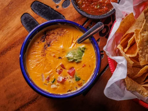 Bowl of Curra's Grill chile con queso topped with guacamole and pico de gallo, beside a basket of tortilla chips and a molcajete of red salsa on a wooden table