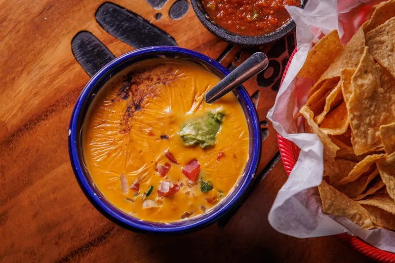 Bowl of Curra's Grill chile con queso topped with guacamole and pico de gallo, beside a basket of tortilla chips and a molcajete of red salsa on a wooden table
