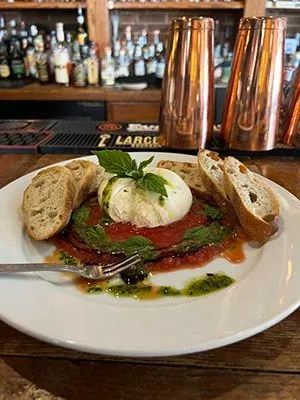 Plate of burrata over San Marzano tomato sauce with basil pesto and sliced baguette at Bourbons Bistro, set on a wooden bar top with copper cocktail shakers and a backbar of bottles behind