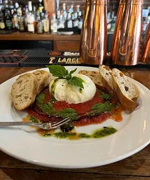 Plate of burrata over San Marzano tomato sauce with basil pesto and sliced baguette at Bourbons Bistro, set on a wooden bar top with copper cocktail shakers and a backbar of bottles behind