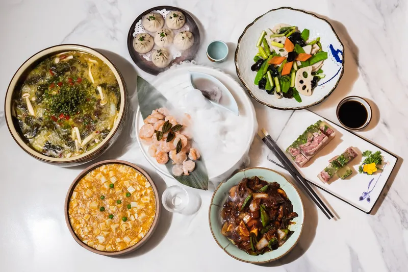 Overhead spread of Shanghainese dishes at CheLi including a steamer of soup dumplings, crab-meat tofu stew, poached shrimp and lotus-root stir-fry