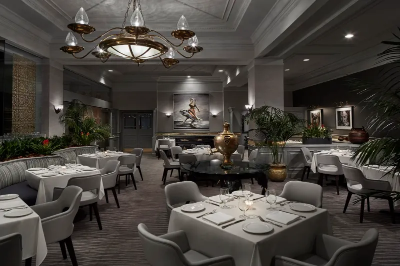 The Bombay Club dining room in Washington, DC, with white-clothed tables, gray armchairs, a brass chandelier, and a brass samovar centerpiece