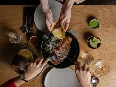Overhead view of Cosme's Pekin duck carnitas in a cast-iron pan with corn tortillas, salsas and lime for tableside tacos