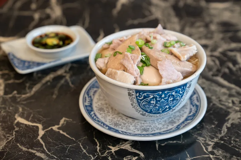 A bowl of Hong Kong-style noodle soup with sliced meats and scallions, served with a dish of chili sauce, at Kenny's Noodle House in Portland