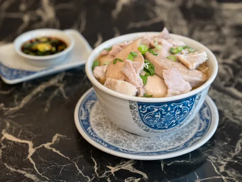A bowl of Hong Kong-style noodle soup with sliced meats and scallions, served with a dish of chili sauce, at Kenny's Noodle House in Portland