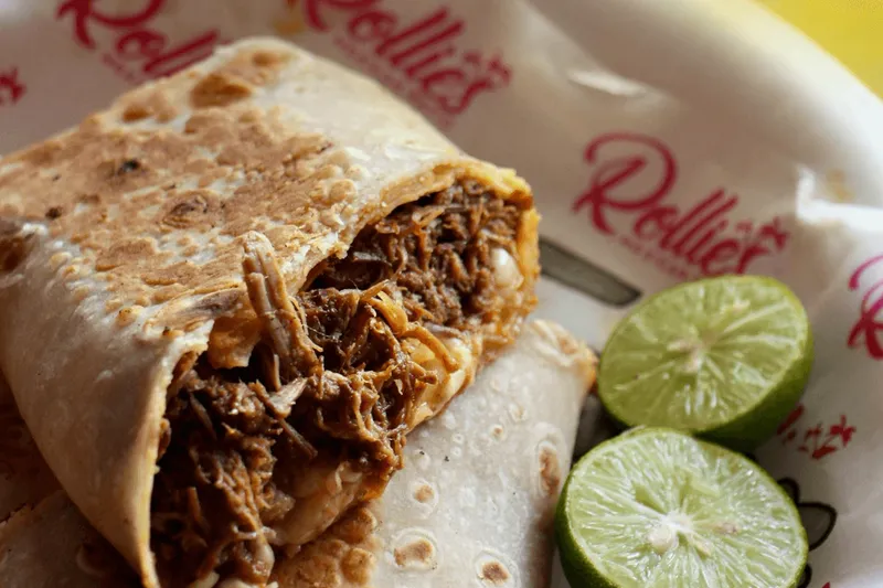 A cut-open birria burrito showing shredded stewed beef and melted cheese in a griddled flour tortilla, served on Rollies Mexican Patio branded paper with two lime halves