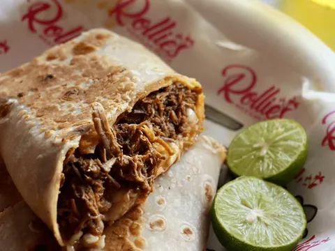A cut-open birria burrito showing shredded stewed beef and melted cheese in a griddled flour tortilla, served on Rollies Mexican Patio branded paper with two lime halves