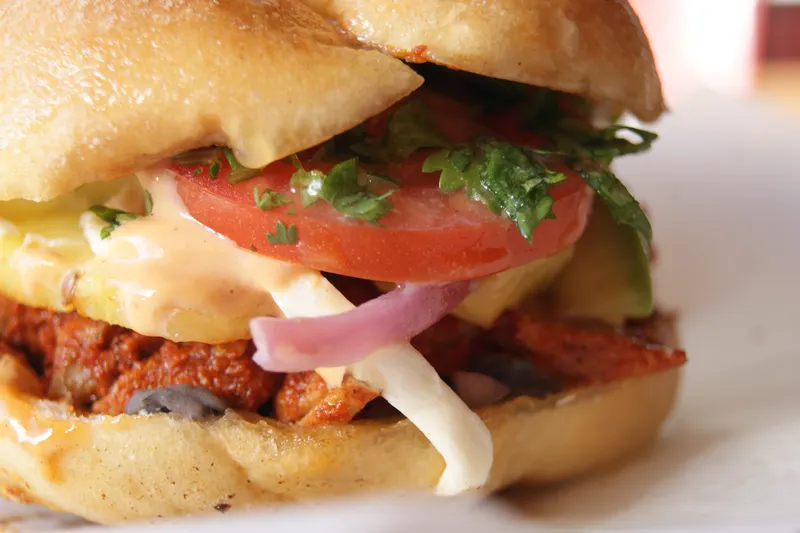 Close-up of a Mexican torta on a telera roll with al pastor pork, melted cheese, tomato, red onion, avocado and cilantro at Tenoch Mexican in Medford