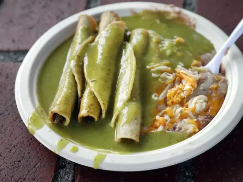 A white plate of Cielito Lindo's rolled beef taquitos smothered in the signature green avocado sauce, with refried beans and shredded cheese, set on an Olvera Street brick surface