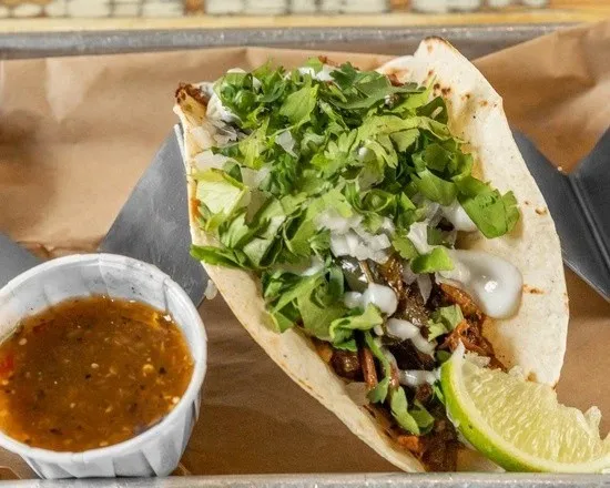 A suadero taco on a flour tortilla topped with cilantro, onion and crema with a lime wedge and a cup of salsa verde, from KC Taco Company in Kansas City