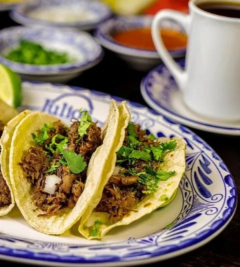 Three barbacoa tacos on soft corn tortillas topped with cilantro and onion, served on a blue-and-white Talavera plate with a cup of consomme at Julio's Cafe Corona