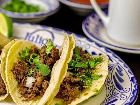 Three barbacoa tacos on soft corn tortillas topped with cilantro and onion, served on a blue-and-white Talavera plate with a cup of consomme at Julio's Cafe Corona