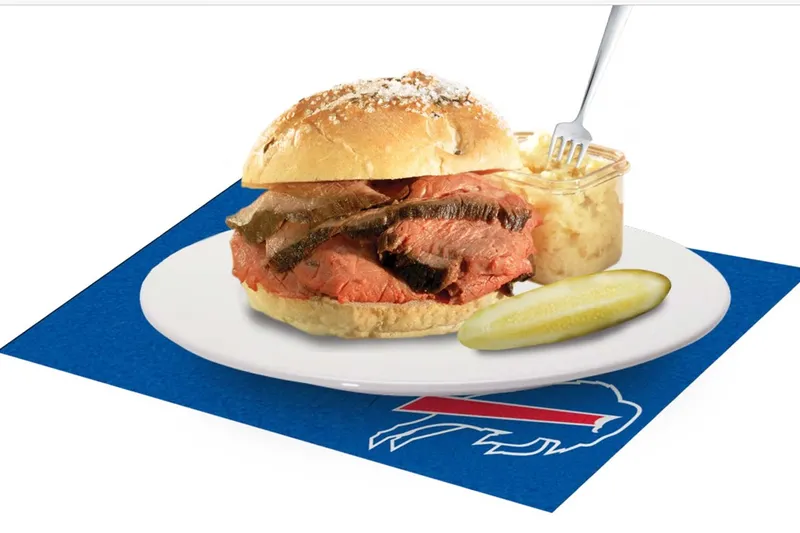 A beef on weck sandwich of carved roast beef on a kummelweck roll with a pickle and side of horseradish at Charlie the Butcher, plated on a Buffalo Bills napkin