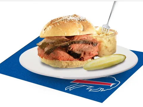 A beef on weck sandwich of carved roast beef on a kummelweck roll with a pickle and side of horseradish at Charlie the Butcher, plated on a Buffalo Bills napkin