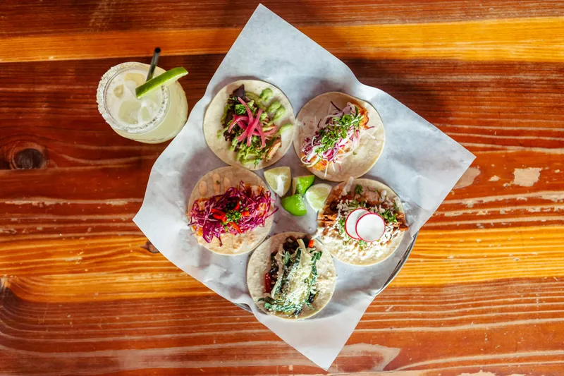 Overhead spread of five Bakersfield street tacos with pickled onion, radish and cilantro alongside a margarita on a dark table