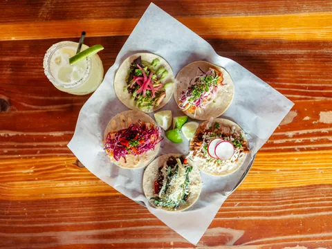 Overhead spread of five Bakersfield street tacos with pickled onion, radish and cilantro alongside a margarita on a dark table