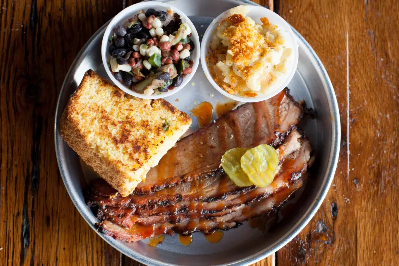 Edley's Bar-B-Que sliced brisket platter with grilled cornbread, mac and cheese, bean salad and pickles on a metal plate