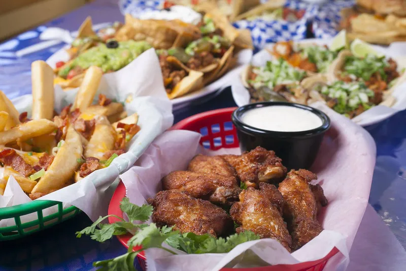 A spread of Cilantro's Mexican Restaurant dishes on a blue table — a basket of fried chicken wings with ranch, bacon-and-cheese loaded fries, guacamole nachos and street tacos