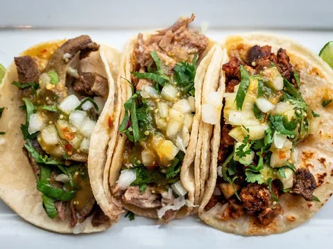 Three El Gallo Giro street tacos on corn tortillas topped with grilled meats, salsa verde, onion and cilantro, with lime wedges