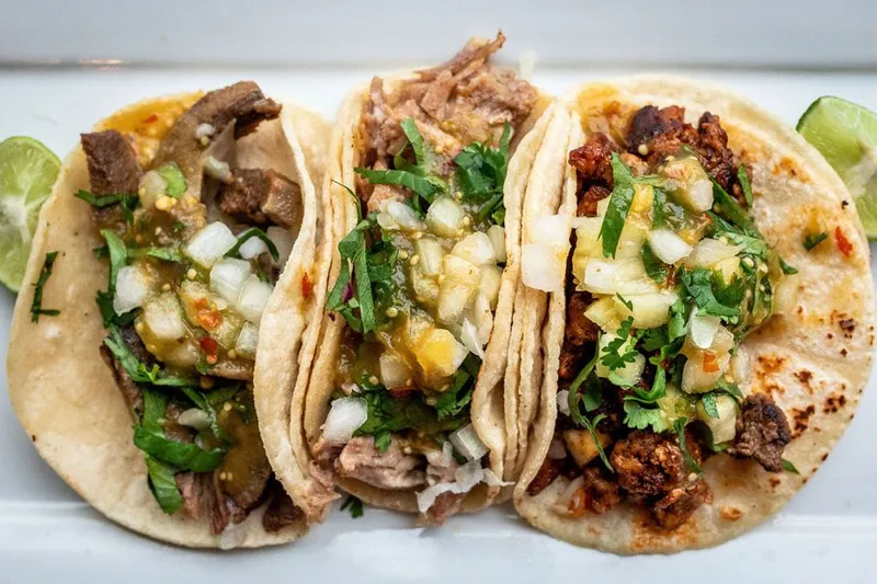 Three El Gallo Giro street tacos on corn tortillas topped with grilled meats, salsa verde, onion and cilantro, with lime wedges