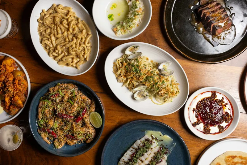 Overhead spread of Kabawa's Caribbean dishes — cacio e pepe-style macaroni, seared steak, clam pasta, pepper-crusted fish, ceviche and chili oil over yogurt — on a wooden table