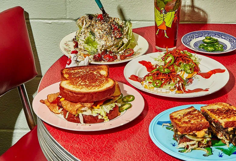 A spread of Turkey and the Wolf dishes — a wedge salad, fried bologna sandwich, loaded fries, and a cocktail — on a red diner table