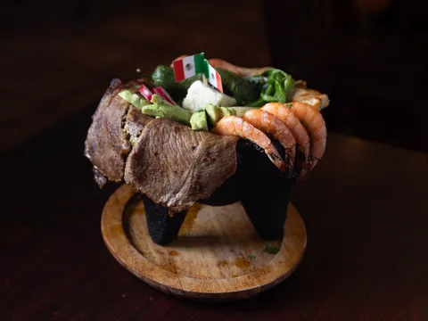 A signature molcajete at Los Molcajetes — a lava-stone bowl on a wooden plate overflowing with grilled steak, shrimp, chicken, avocado and grilled peppers, topped with two small Mexican flag picks