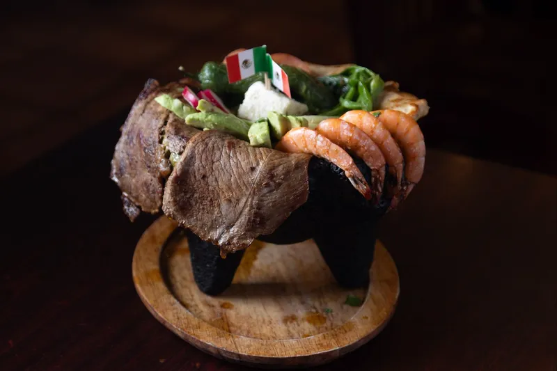 A signature molcajete at Los Molcajetes — a lava-stone bowl on a wooden plate overflowing with grilled steak, shrimp, chicken, avocado and grilled peppers, topped with two small Mexican flag picks