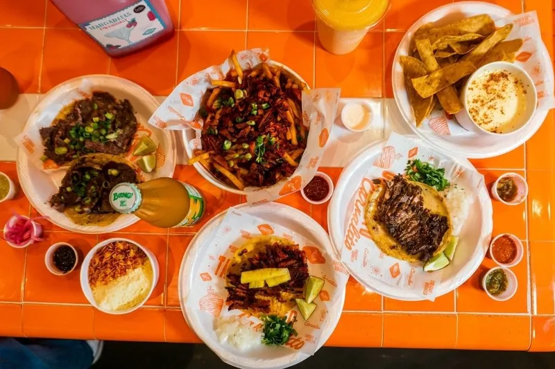 An overhead spread of Taquerias Carnalito dishes on an orange-tiled table — carne asada tacos, al pastor tacos, loaded Desmadre fries, tortilla chips with queso, elote, salsas and a Boing bottle