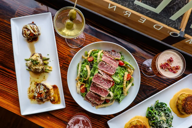 Overhead spread of dishes at ML Steak Modern Chophouse, including a seared tuna salad, filet medallions, a crab cake and cocktails on a wood table