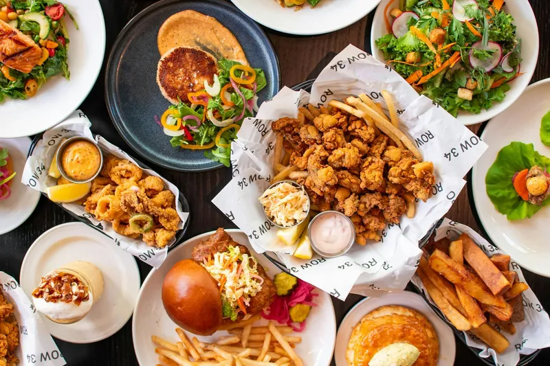 Overhead spread of Row 34 seafood dishes including fried clams, calamari, salads, a fried fish sandwich and fries on ROW 34 branded paper