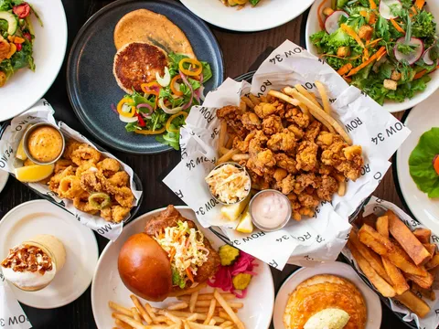 Overhead spread of Row 34 seafood dishes including fried clams, calamari, salads, a fried fish sandwich and fries on ROW 34 branded paper