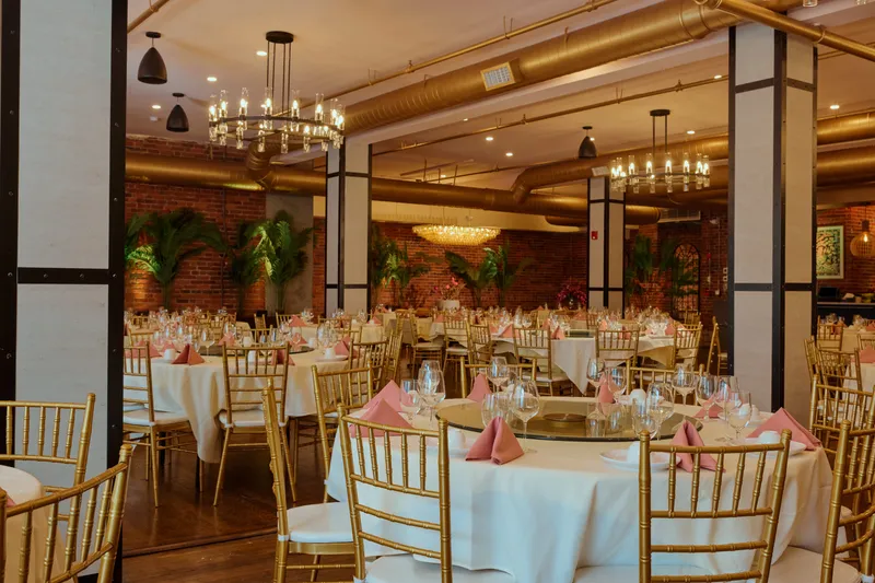 China Pearl's chandelier-lit banquet dining hall in Boston Chinatown, with round tables in white linens, gold Chiavari chairs, pink napkins, and exposed-brick walls