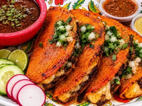 A plate of Taqueria Jalisco quesabirria tacos with crispy orange-tinted tortillas, melted cheese and cilantro, served with a cup of birria consommé, red and green salsas, lime, cucumber and radish