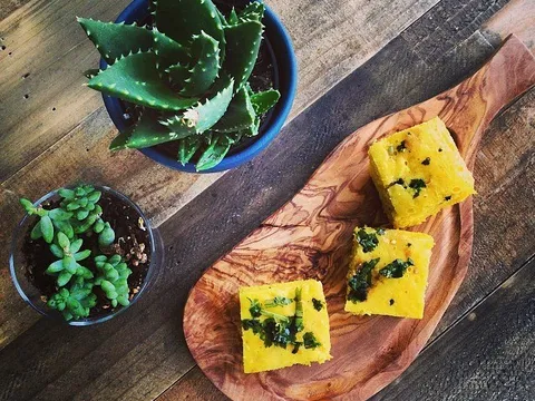 Squares of khaman dhokla topped with cilantro and green chili, served on a wooden board beside potted succulents at Usha Foods in Floral Park