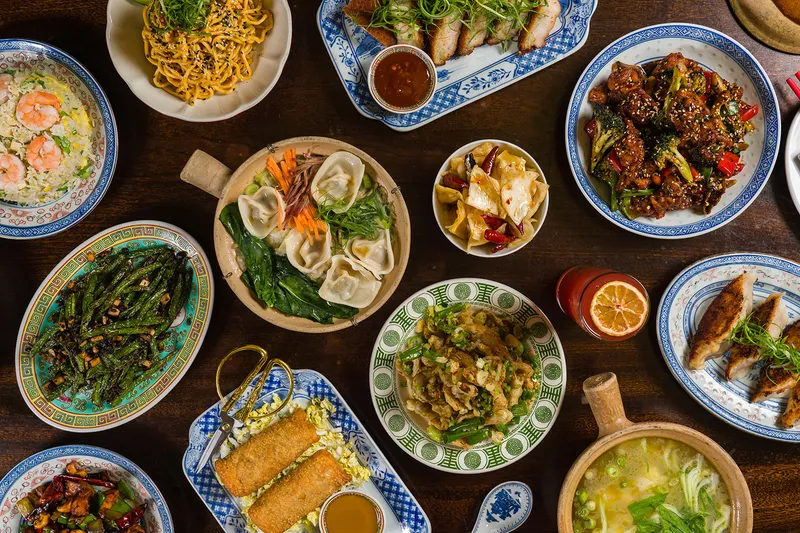 Overhead spread of dishes at Kings County Imperial including dumplings, noodles, dry-fried long beans, fried rice, spring rolls, and a claypot soup
