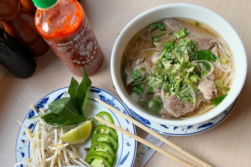 Bowl of beef phở with rice noodles, sliced beef, cilantro and scallions in a blue-and-white bowl at Pho Lang Thang, with a side plate of bean sprouts, Thai basil, lime and sliced jalapeños